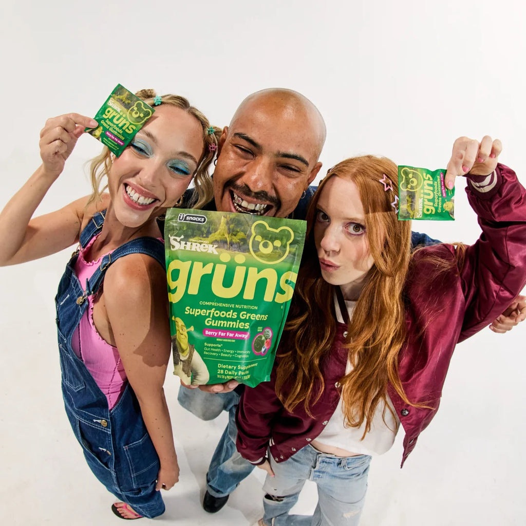 Three people posing playfully together, each holding up a green Shrek Grüns Superfoods Greens Gummies package, with the large main pack labeled Berry Far Far Away held in the center.