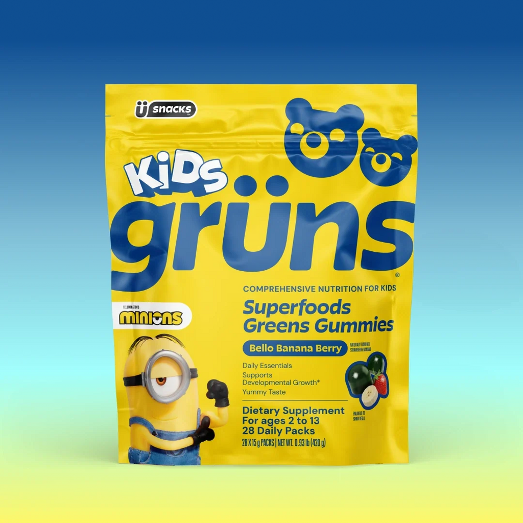 A yellow bag of Kids Grüns Superfoods Greens Gummies in "Bello Banana Berry" flavor features a Minions character on the front, labeled as a dietary supplement for ages 2 to 13 with 28 daily packs, set against a blue-to-yellow gradient background.