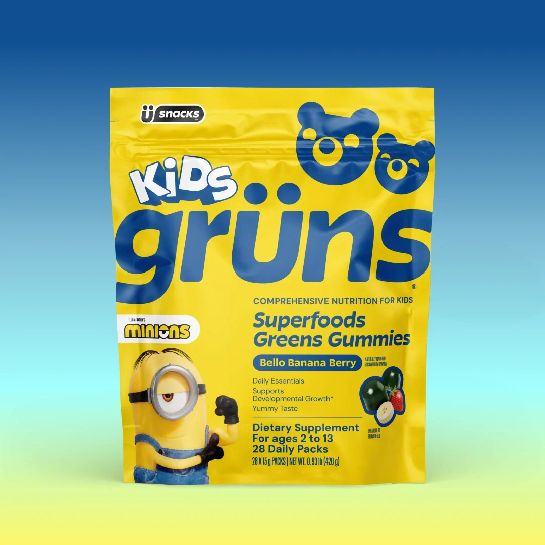 A yellow bag of Kids Grüns Superfoods Greens Gummies in "Bello Banana Berry" flavor features a Minions character on the front, labeled as a dietary supplement for ages 2 to 13 with 28 daily packs, set against a blue-to-yellow gradient background.