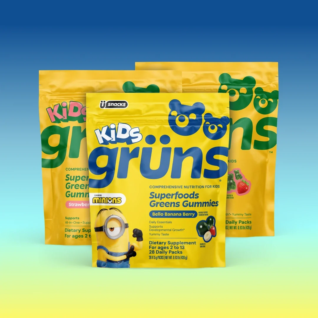 Three yellow Kids Grüns Superfoods Greens Gummies packets in Strawberry and Bello Banana Berry flavors, the front packet featuring Minions branding, on a blue-to-yellow gradient background