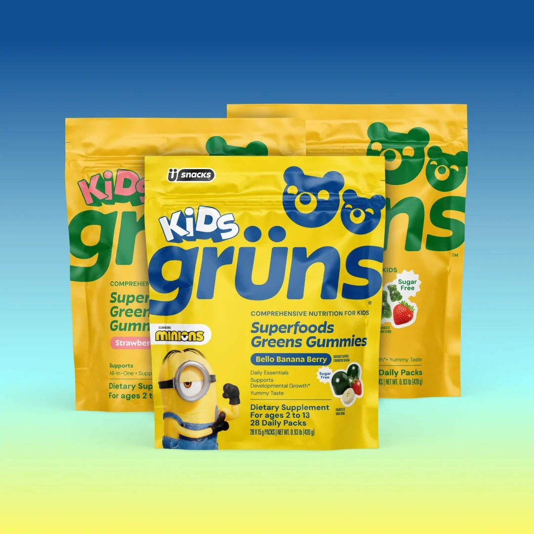 Three yellow Kids Grüns Superfoods Greens Gummies packets in Strawberry and Bello Banana Berry flavors, the front packet featuring Minions branding, on a blue-to-yellow gradient background