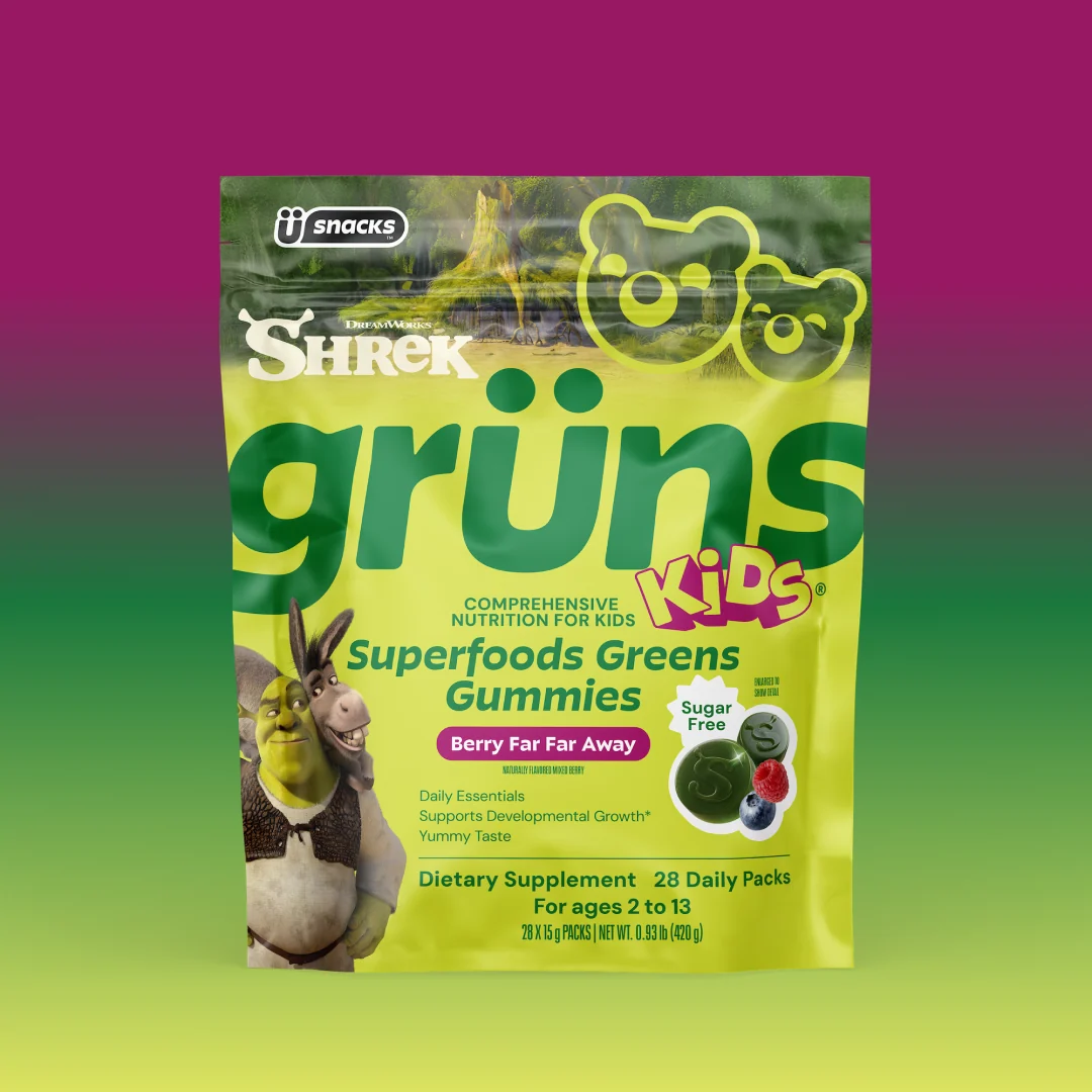 Yellow-green Shrek Kids Grüns Superfoods Greens Gummies package featuring Shrek and Donkey, labeled Berry Far Far Away flavor with a Sugar Free badge, listing Daily Essentials, Supports Developmental Growth, Yummy Taste, Dietary Supplement, 28 Daily Packs, for ages 2 to 13, on a pink-to-green gradient background.